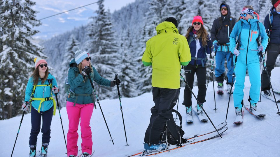 Prowadzący w limonkowej kurtce objaśnia ćwiczenia grupie na nartach skiturowych.