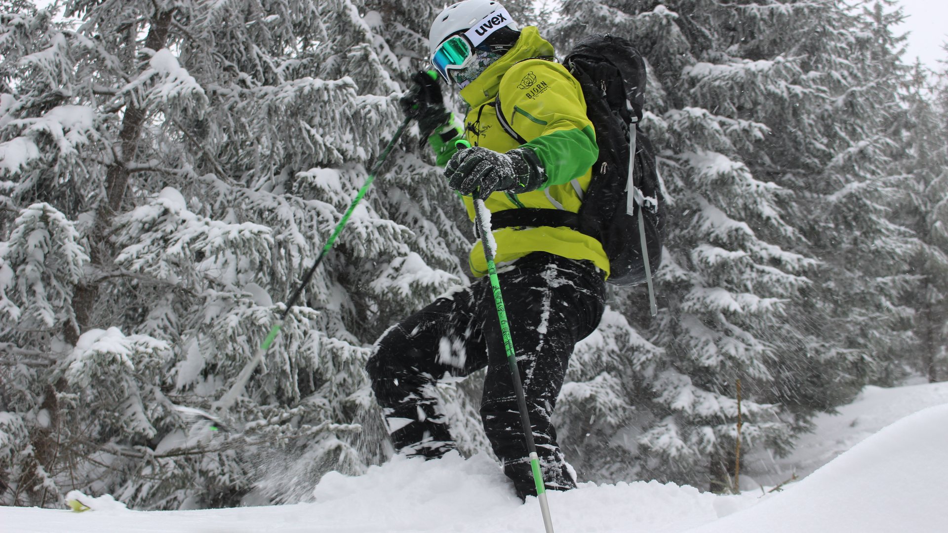 Skiturowiec w zielonej kurtce wspina się przez śnieg z oznaczeniem BJØRN TO SKI na ramieniu.